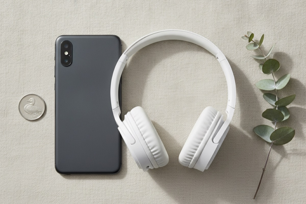 Flat-lay of a smartphone and white over-ear headphones with a Canadian coin and a eucalyptus sprig on a soft cream linen background